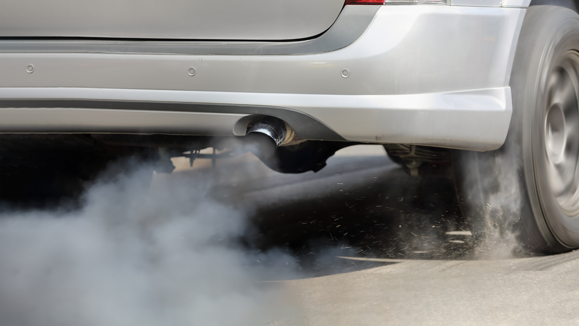 Close-up of a silver car’s rear bumper and exhaust pipe emitting white smoke while driving.
