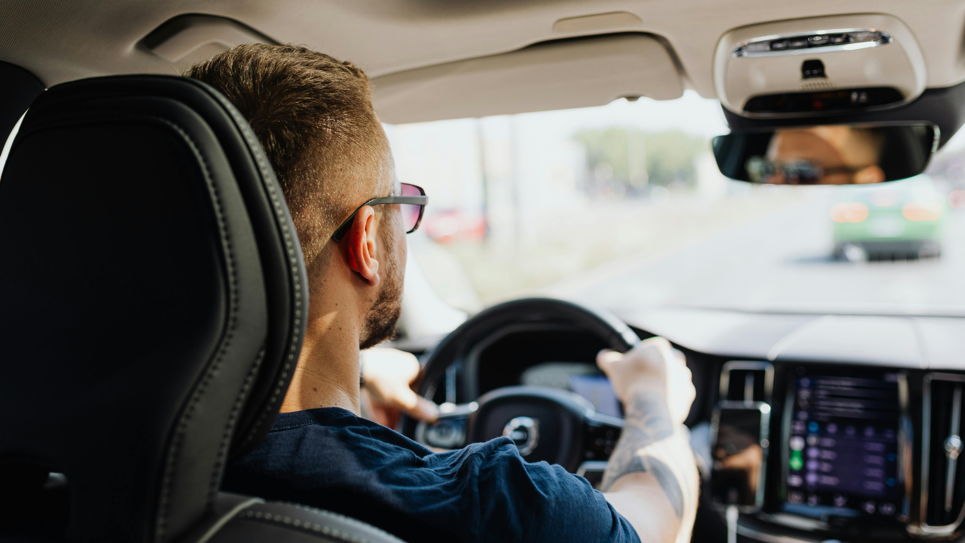 Driver steering a car, seen from the back seat, with a lit dashboard and road ahead.