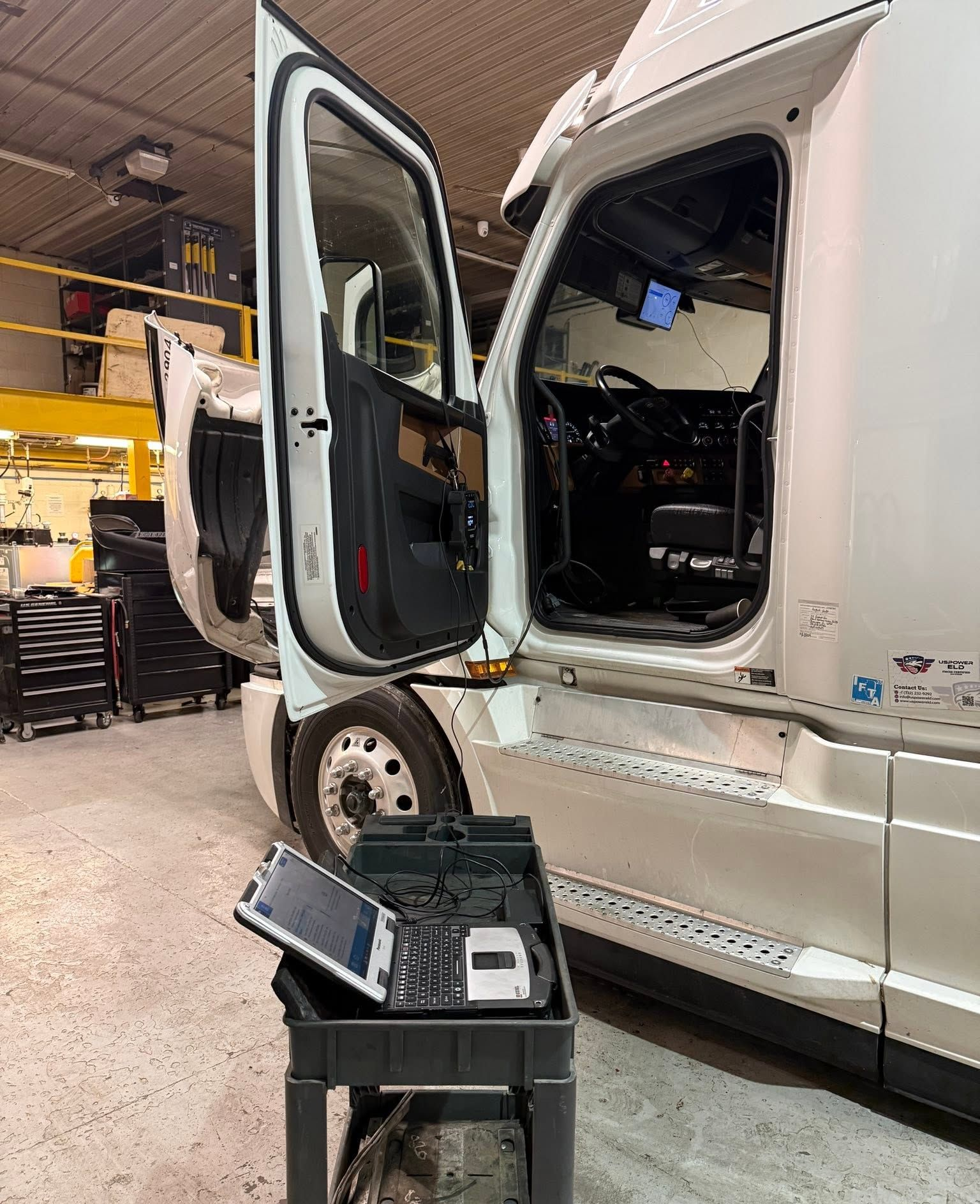 White semi-truck in a garage with driver door open and diagnostic equipment beside it