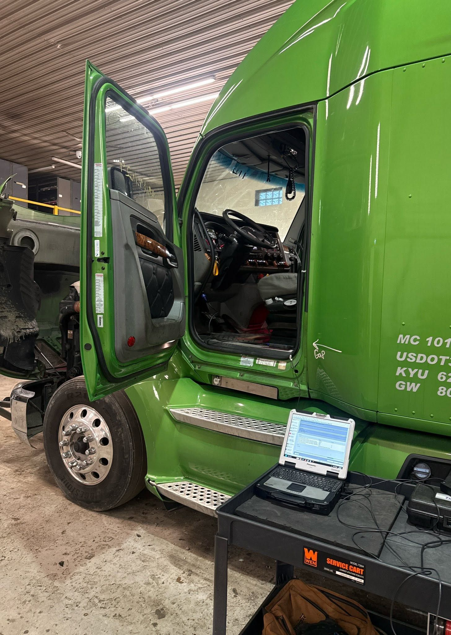 Green semi-truck cab with open driver door in a garage or showroom, black equipment cart beside it.