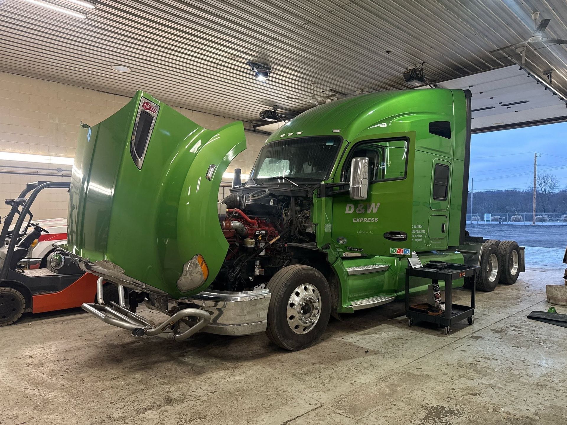 Green semi-truck with hood raised in a garage, showing front damage and exposed engine parts.