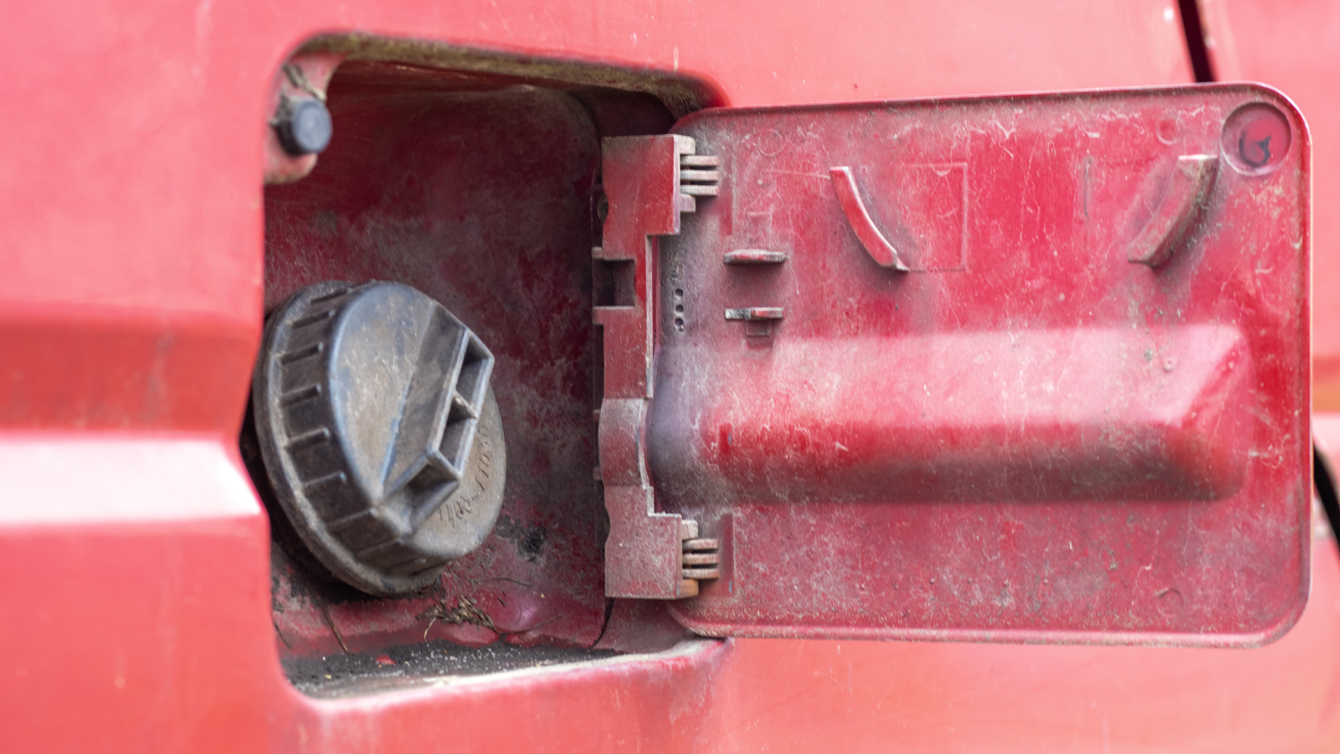 Red plastic fuel cap open on a red vehicle body, showing the fuel filler compartment.
