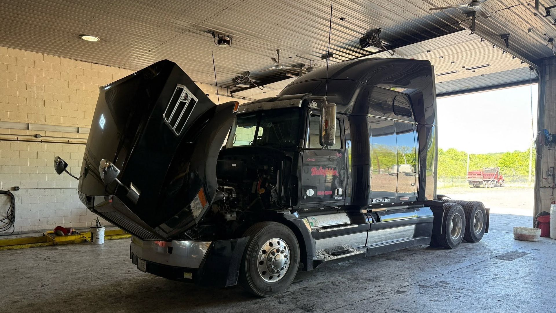 Damaged black semi-truck inside a garage, hood open and cab tilted after a crash.