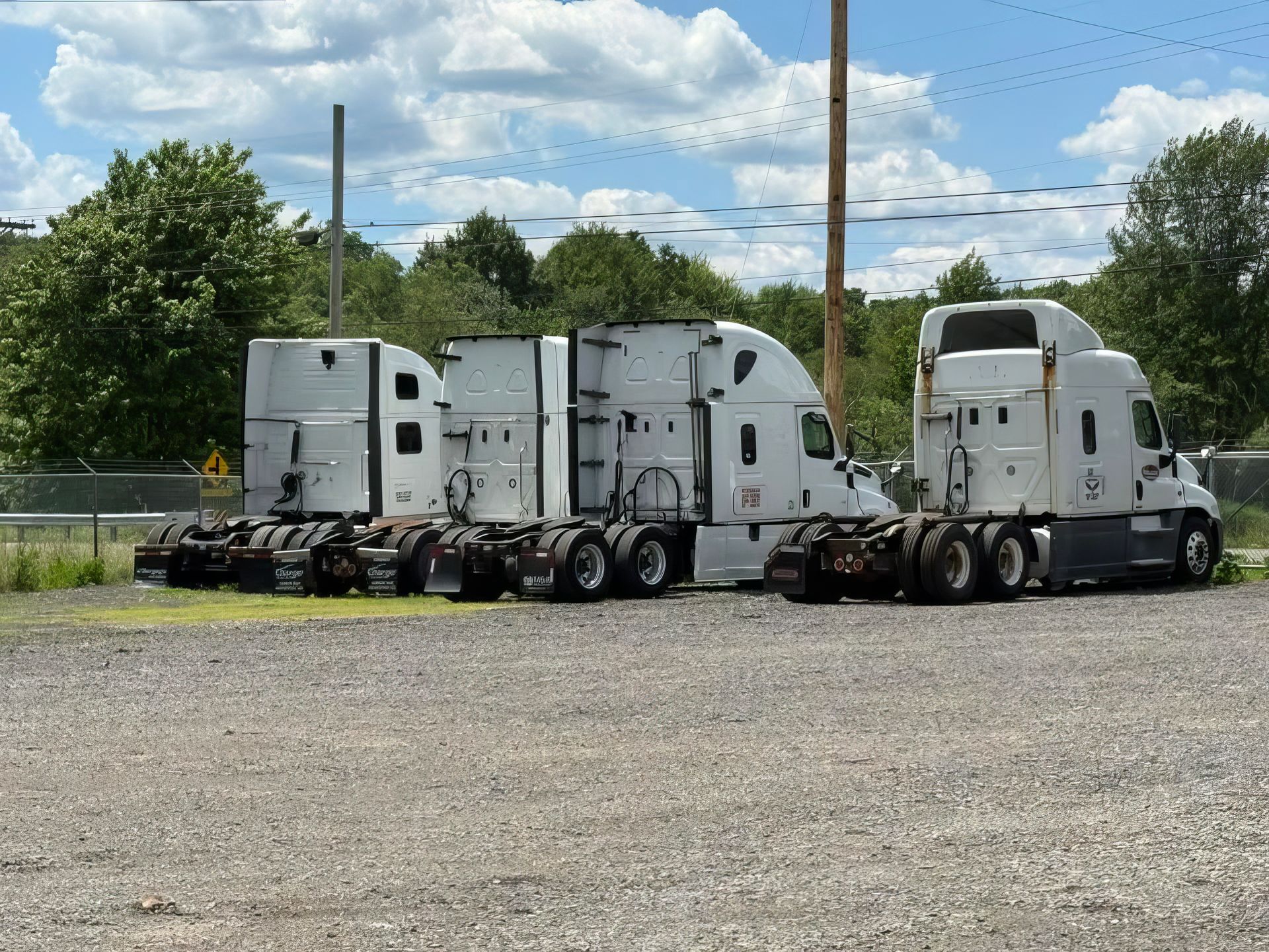Three white semi-truck cabs parked side by side on a gravel lot under a cloudy blue sky