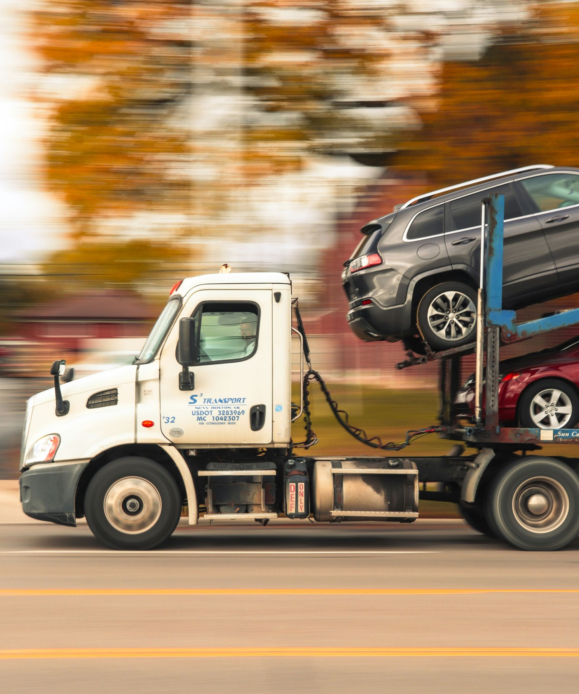 Tow truck carrying a gray SUV on a flatbed, speeding past blurred autumn trees