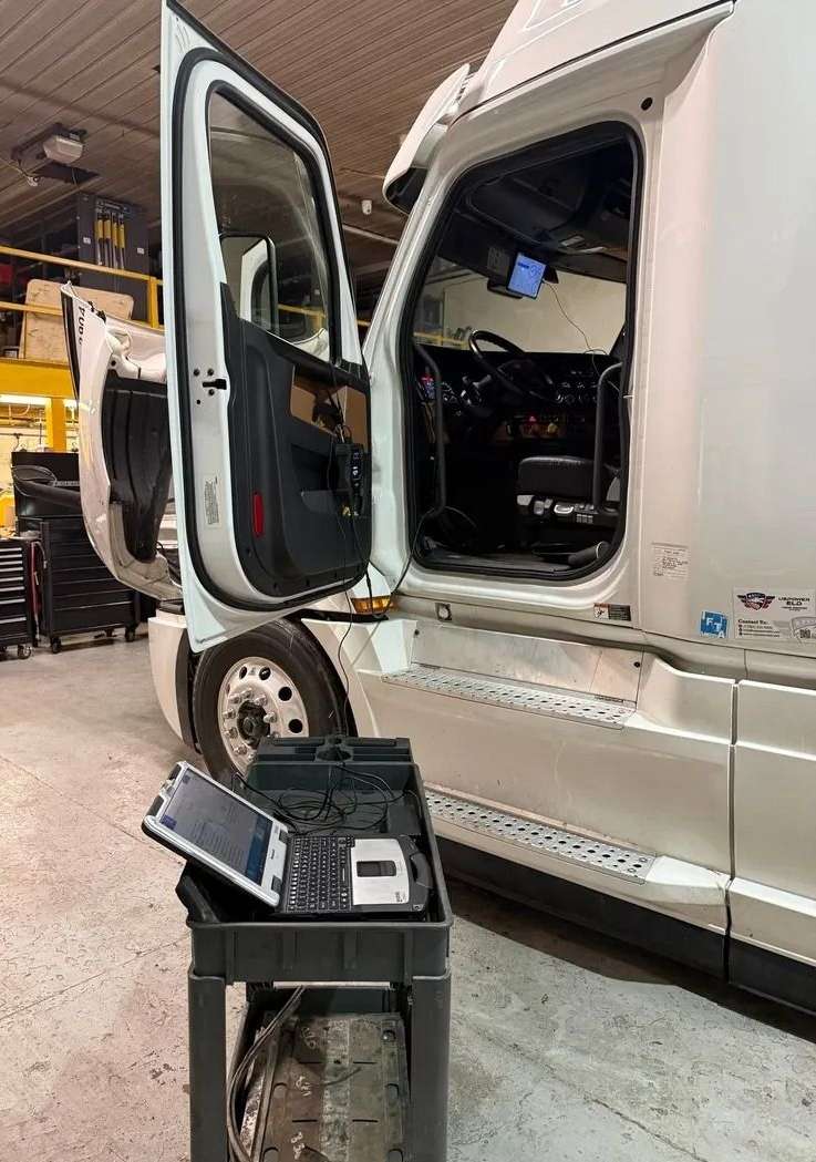 White semi-truck in a garage with open driver door and exposed cab interior, shown beside a tool cart.