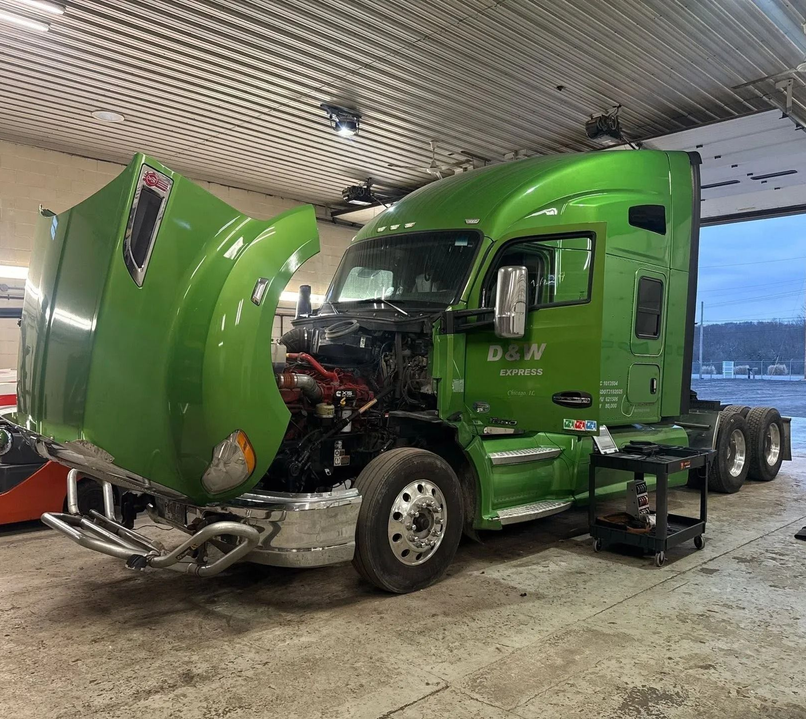 Green semi-truck in a garage with the hood open and front-end damage visible