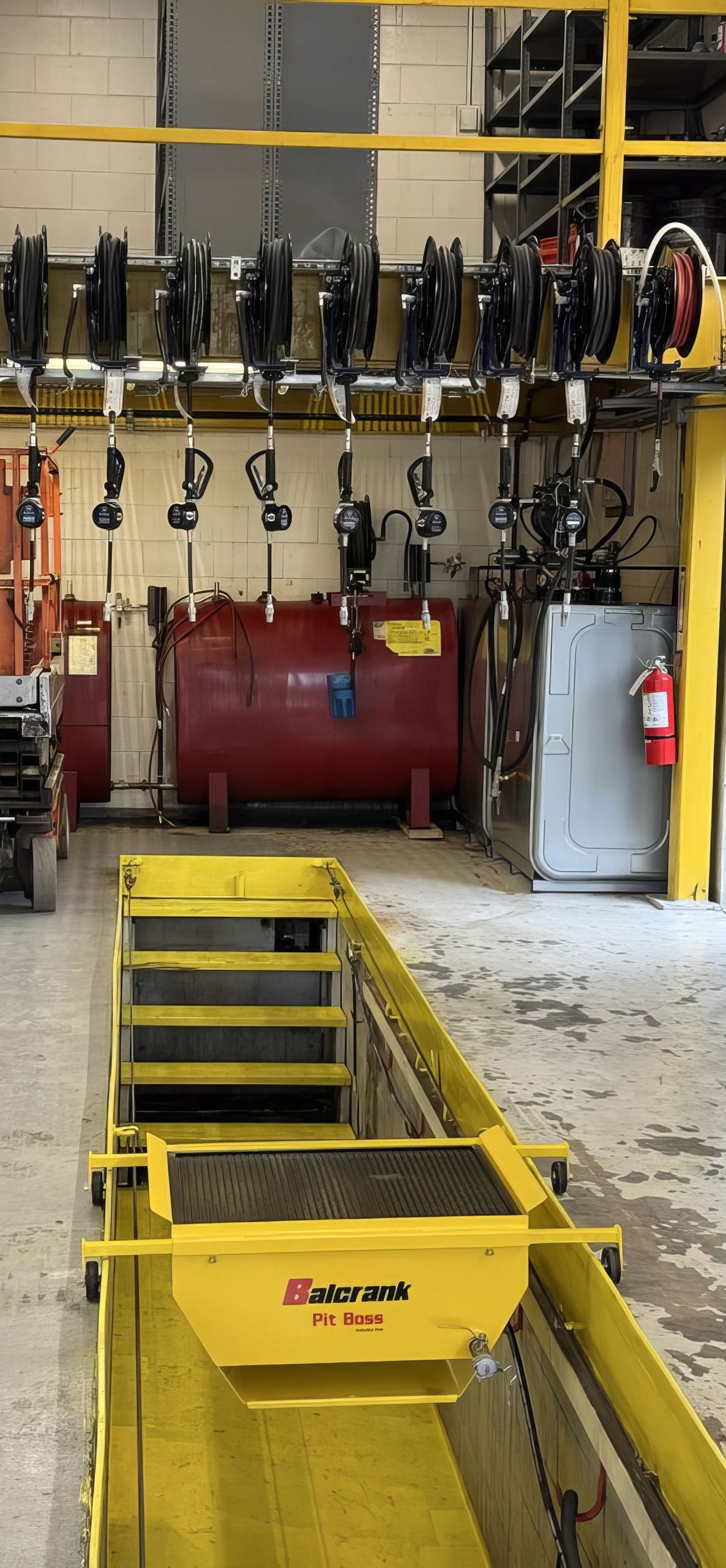 Industrial workshop with yellow machinery lift, hanging tools, and red equipment against a tiled wall