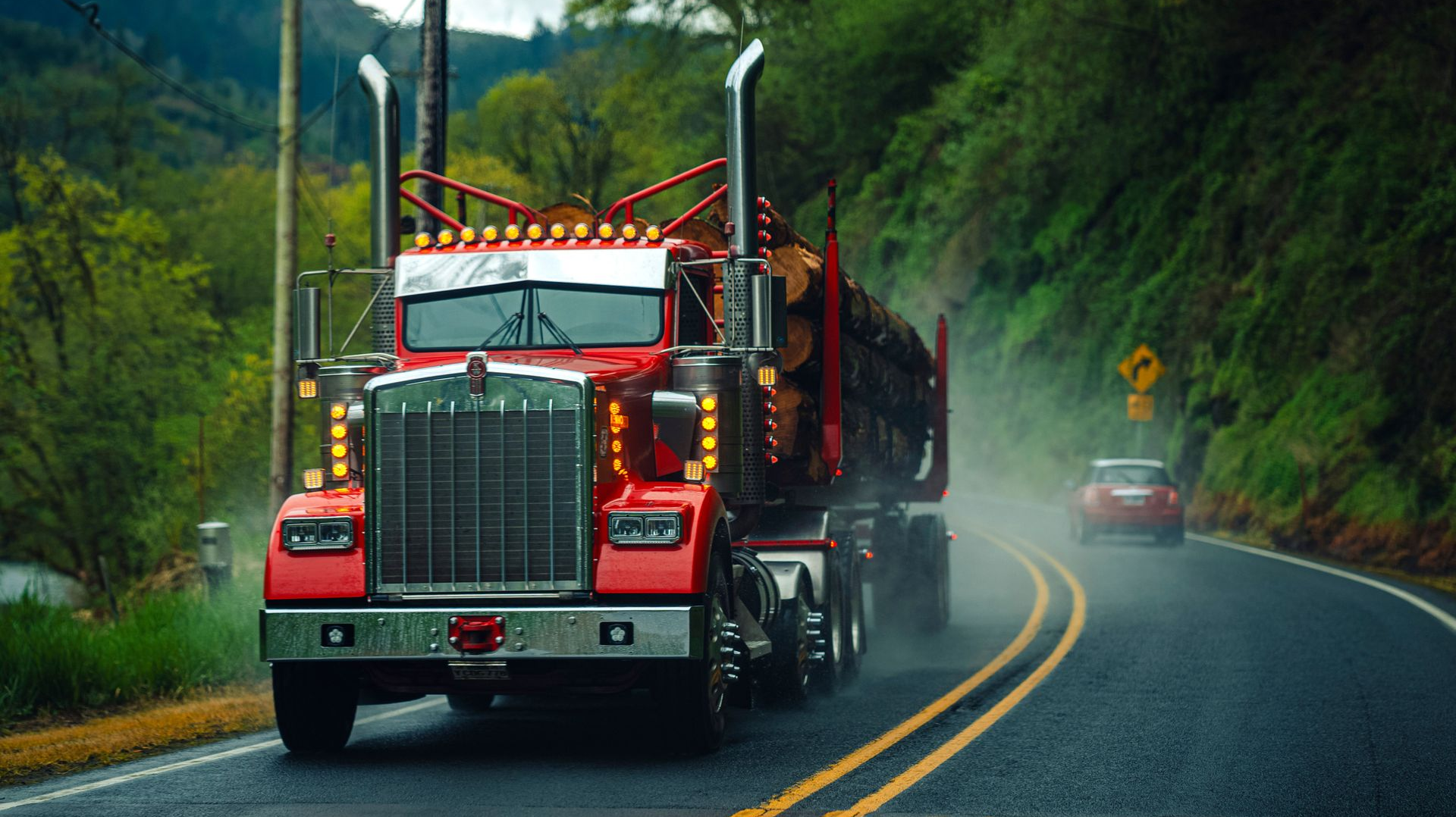 Red logging truck driving on a wet winding mountain road in mist, with a car behind it