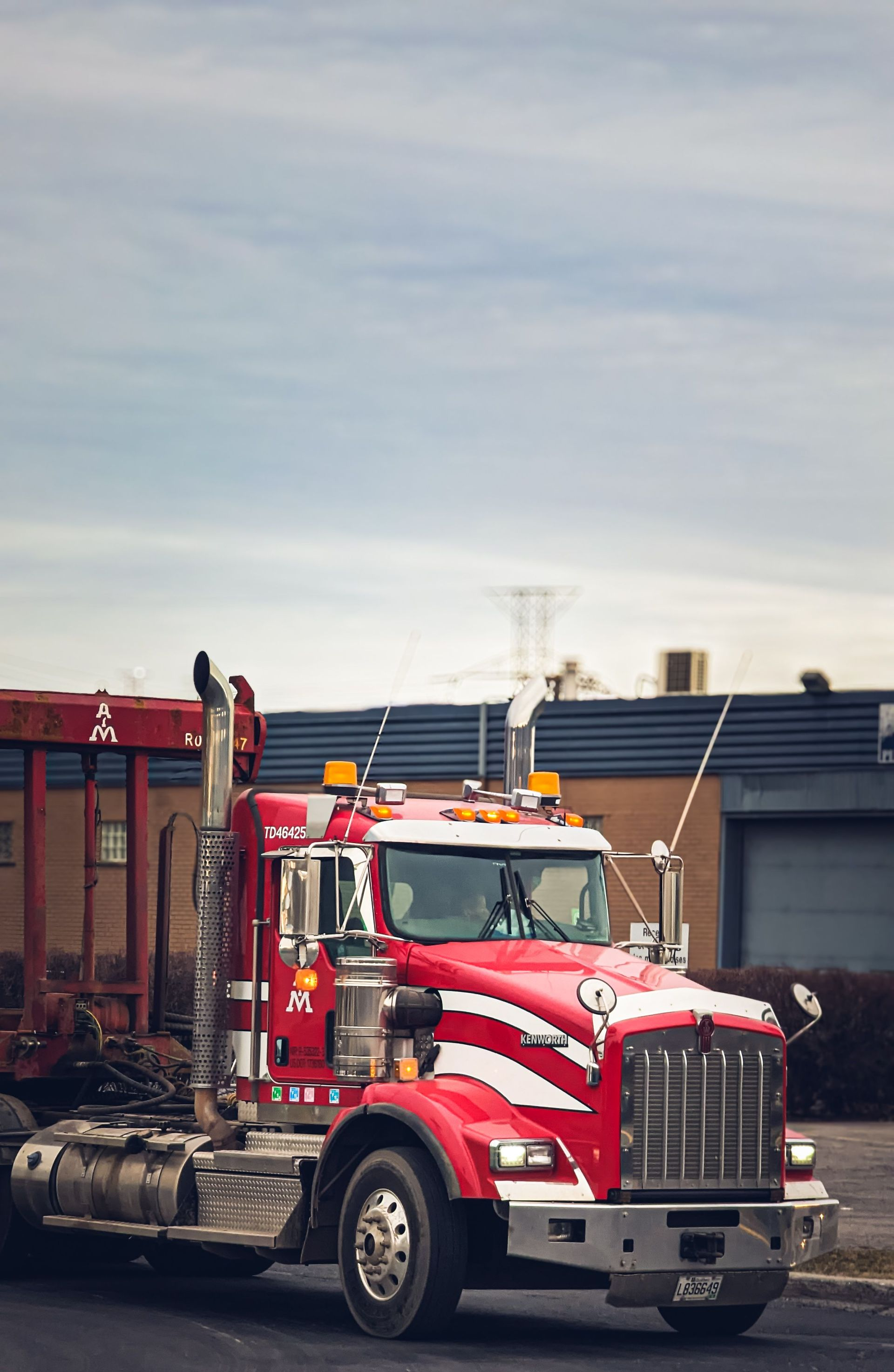 Red semi-truck with trailer parked near industrial buildings under a cloudy sky
