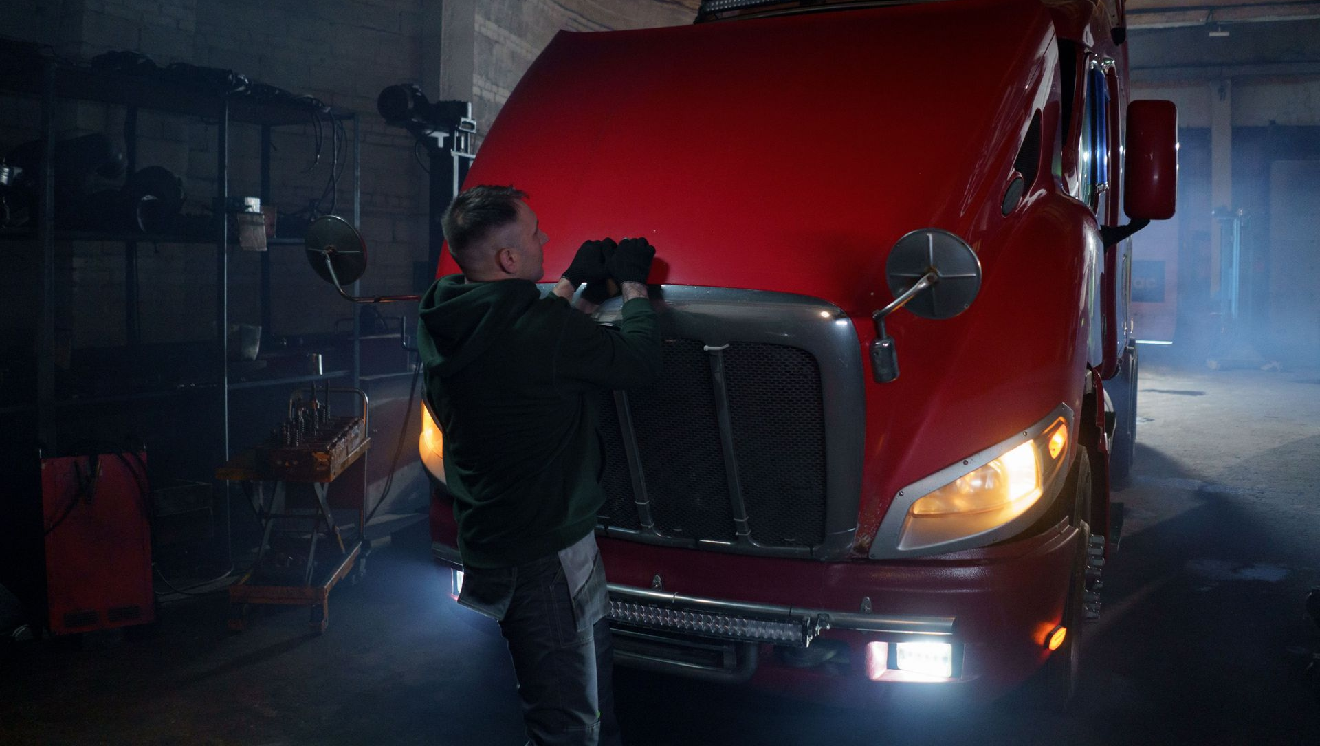 Person inspecting the front of a red semi-truck in a dim warehouse at night