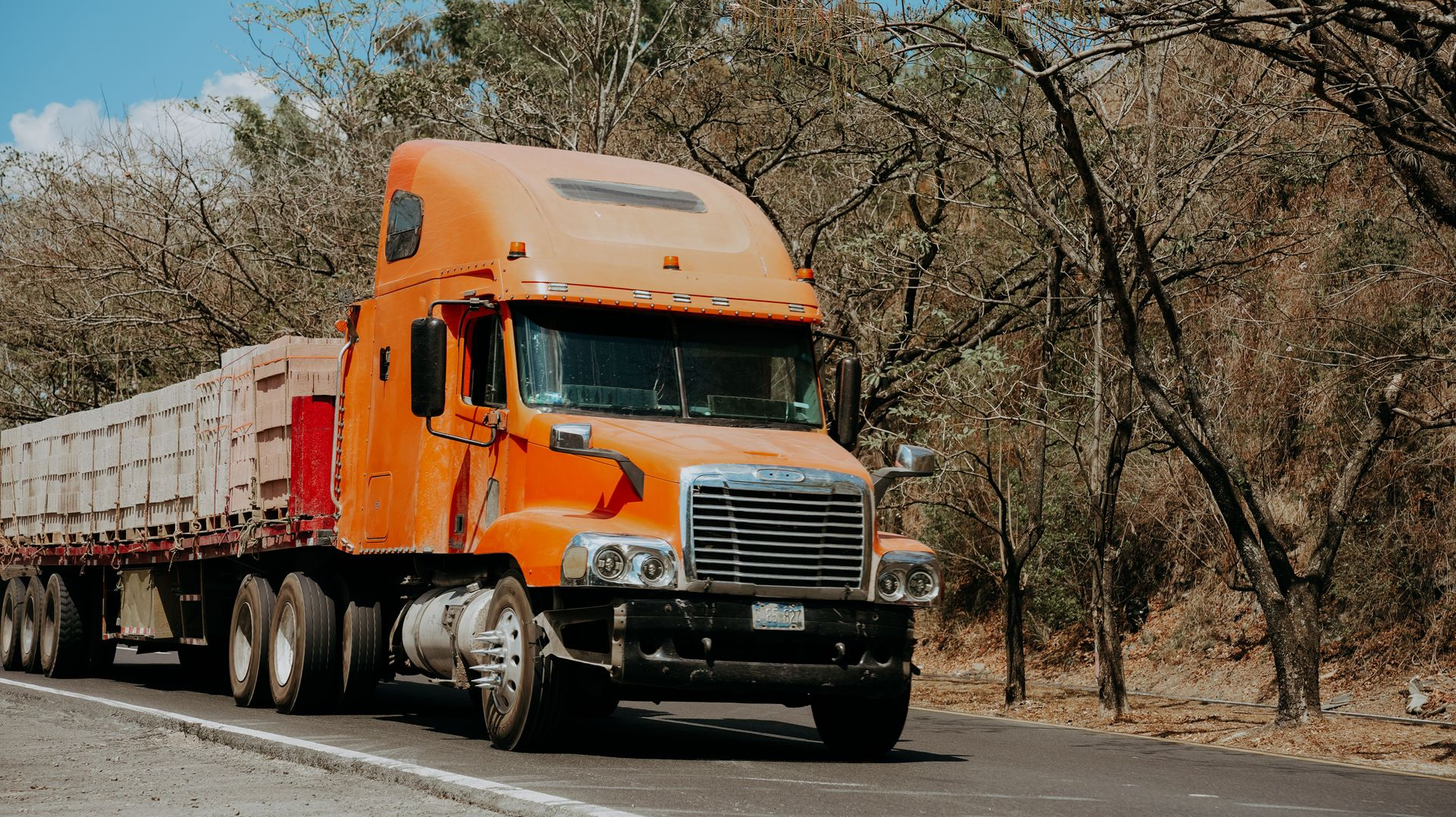 Orange semi-truck hauling a long trailer on a rural road with dry trees