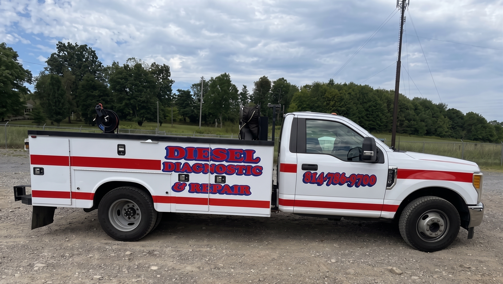 White service pickup truck with red lettering parked on gravel beside trees and a cloudy sky