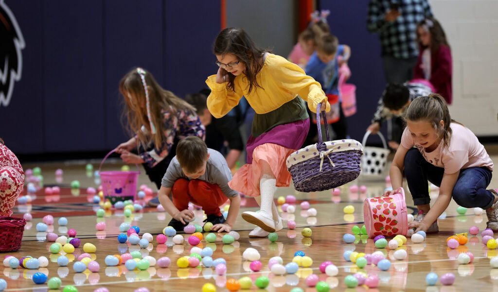 Children kneel on a gym floor, picking up colorful plastic eggs scattered across the floor.