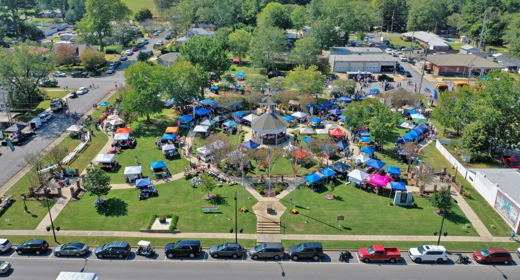 Aerial view of a neighborhood park event with colorful tents, crowds, and parked cars along the street.