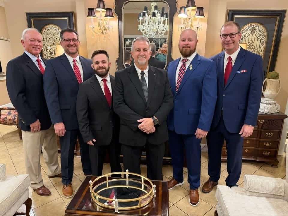 Six men in suits posing in a formal living room beside a glass coffee table