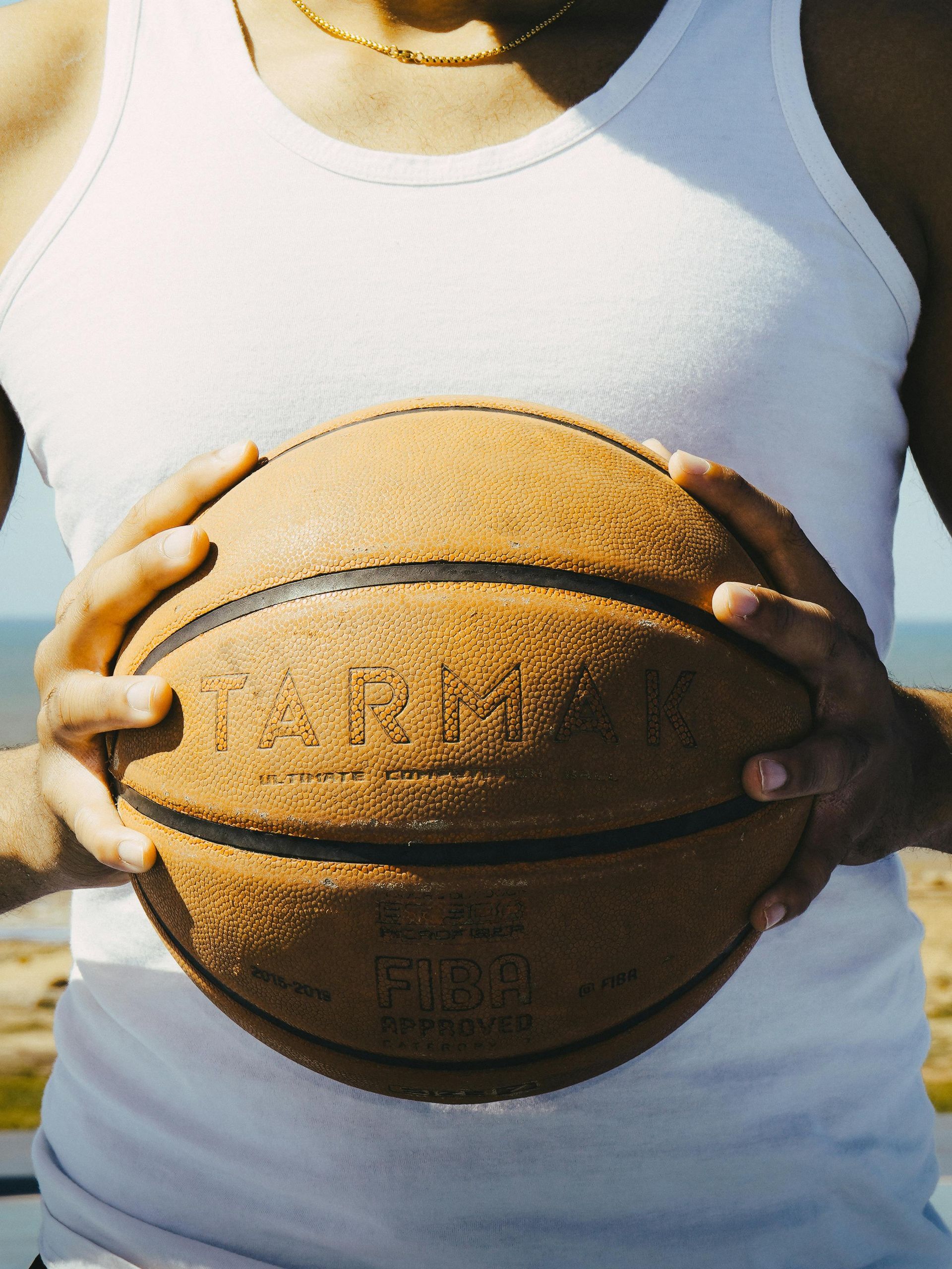 Hands holding a basketball against a white tank top outdoors