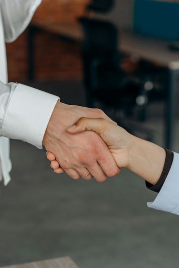 Two people shaking hands in an office setting, one in a white sleeve and one in a light blue sleeve.