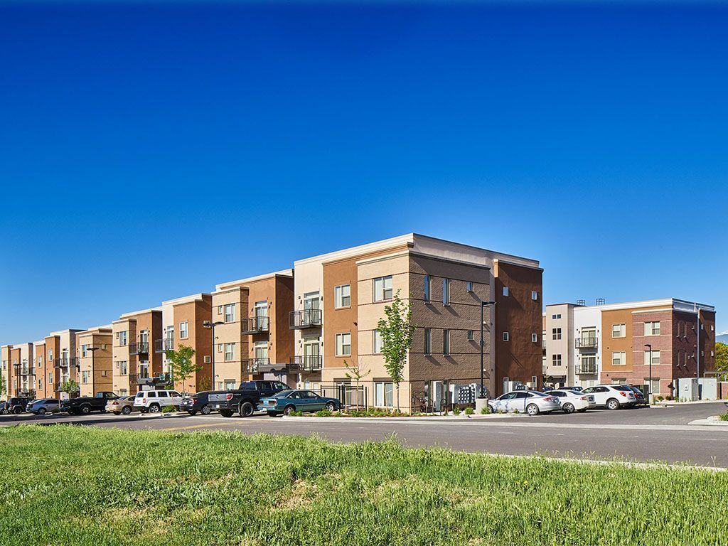 A multi-story apartment complex with tan and brown brick siding sits under a clear blue sky, with cars parked in front.