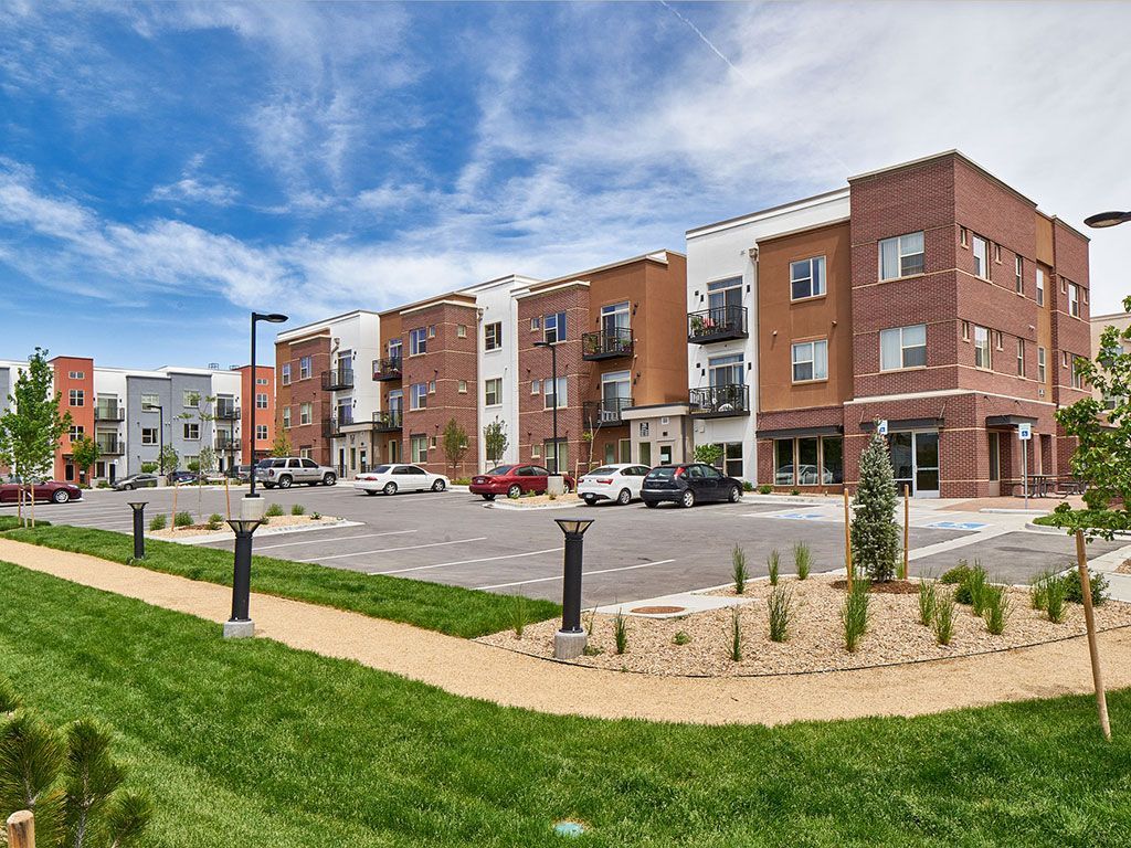 A multi-story apartment complex with brick and white stucco exteriors sits behind a parking lot under a blue sky.