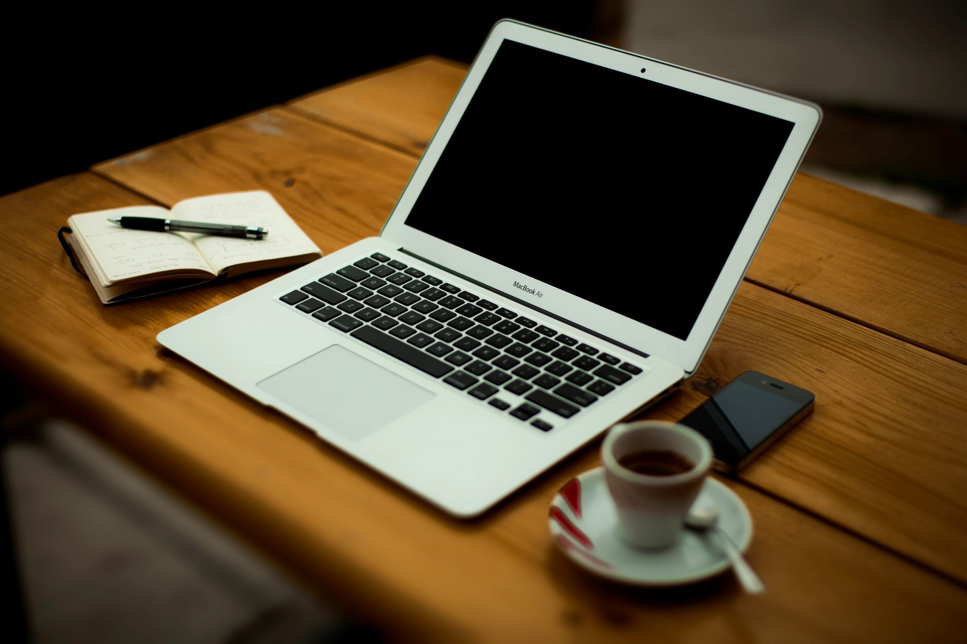 A silver laptop, a notebook with a pen, a smartphone, and a cup of coffee on a wooden table.
