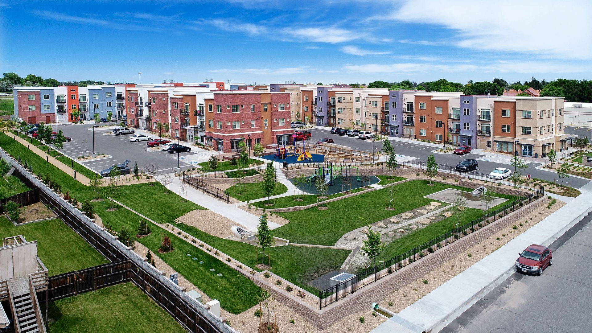 An aerial view of a multi-story apartment complex with a central green courtyard and a paved parking lot on a sunny day.