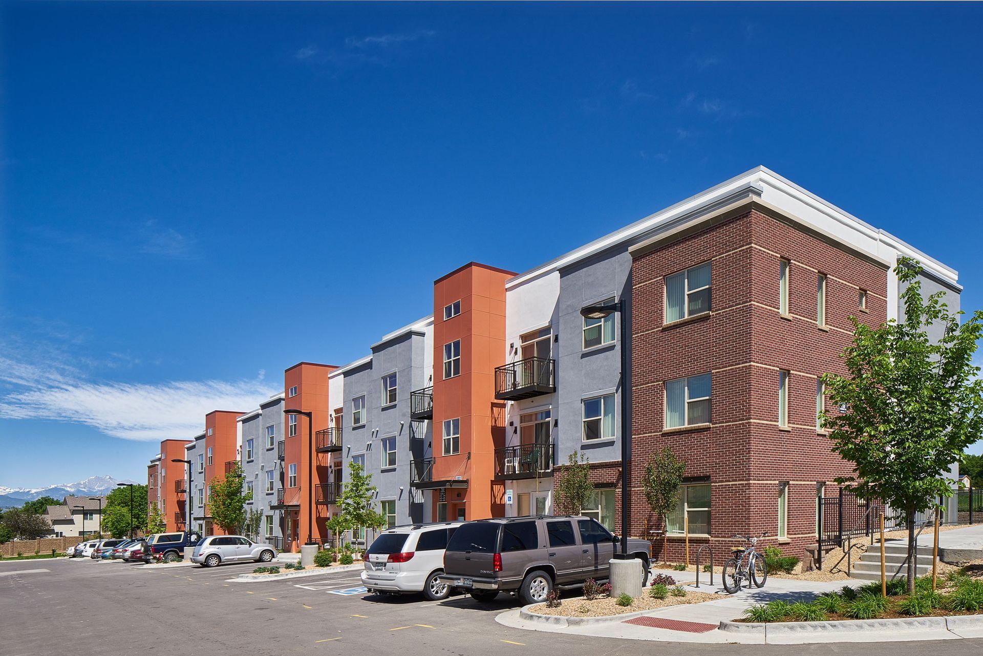 A modern, multi-story apartment complex with brick and grey facades, surrounded by a parking lot and clear blue sky.