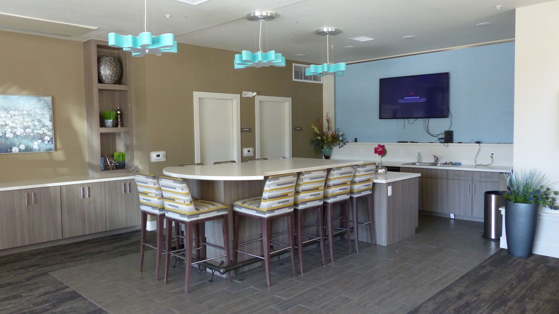 A modern community room featuring a curved bar with patterned stools, light wood cabinets, and three turquoise lights.