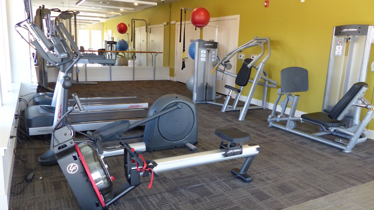 A gym room with treadmills, a rowing machine, and weight machines against a yellow wall on a carpeted floor.