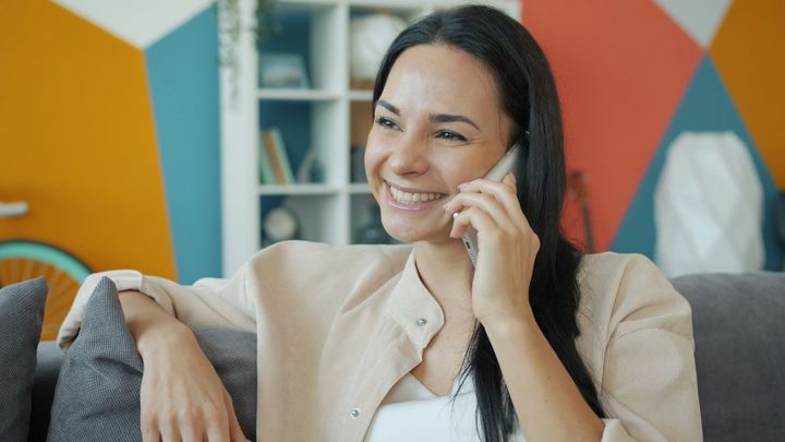A smiling person talks on a smartphone while sitting on a gray couch in a room with a colorful geometric wall.