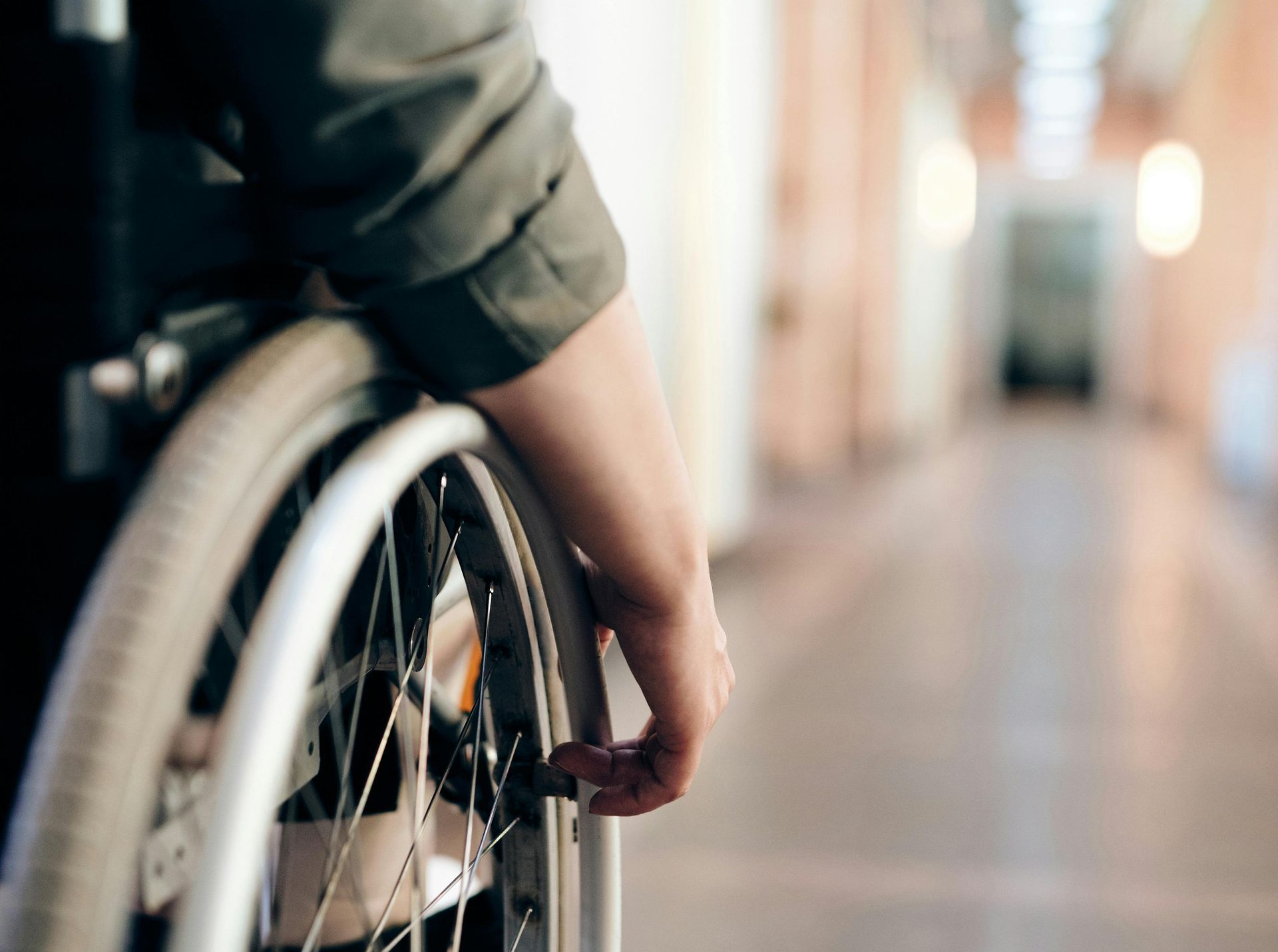 Die Hand einer Person auf einem Rollstuhlrad in einem hell erleuchteten Flur, verschwommener Hintergrund.