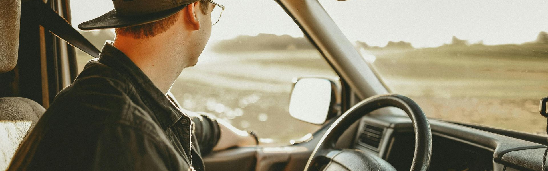 Ein Mann fährt Auto und blickt auf ein Feld. Er trägt einen Hut und eine Brille.