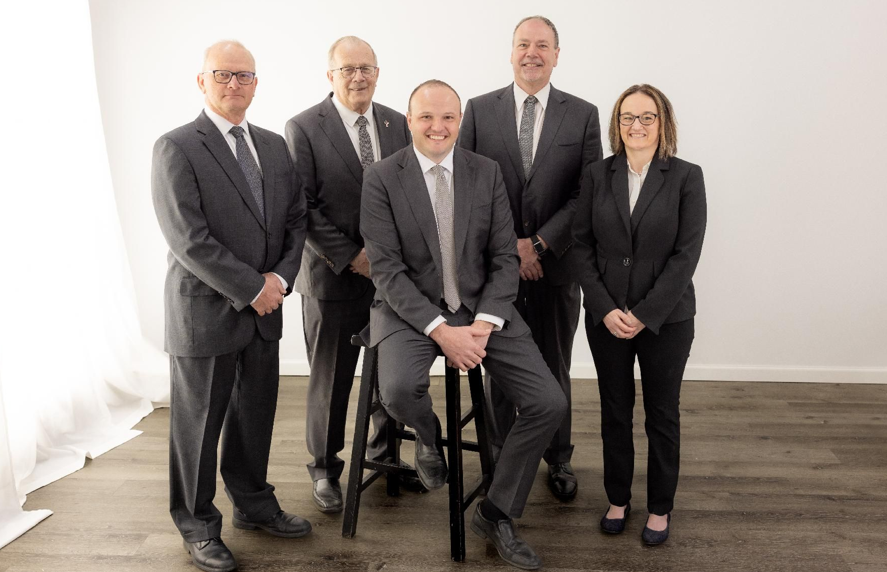 Five professionals in dark suits posing in a bright office, one seated and four standing.
