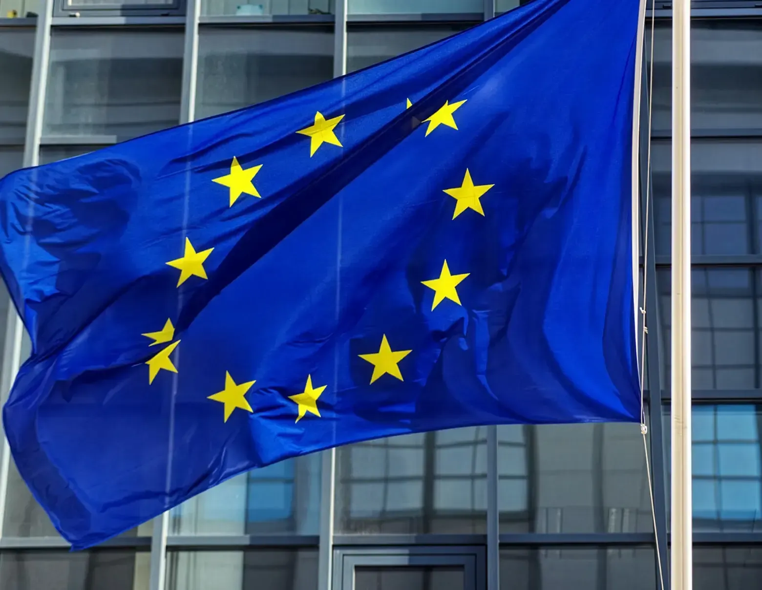 Bandeira azul da União Europeia com estrelas amarelas tremulando em frente a um moderno edifício de vidro.