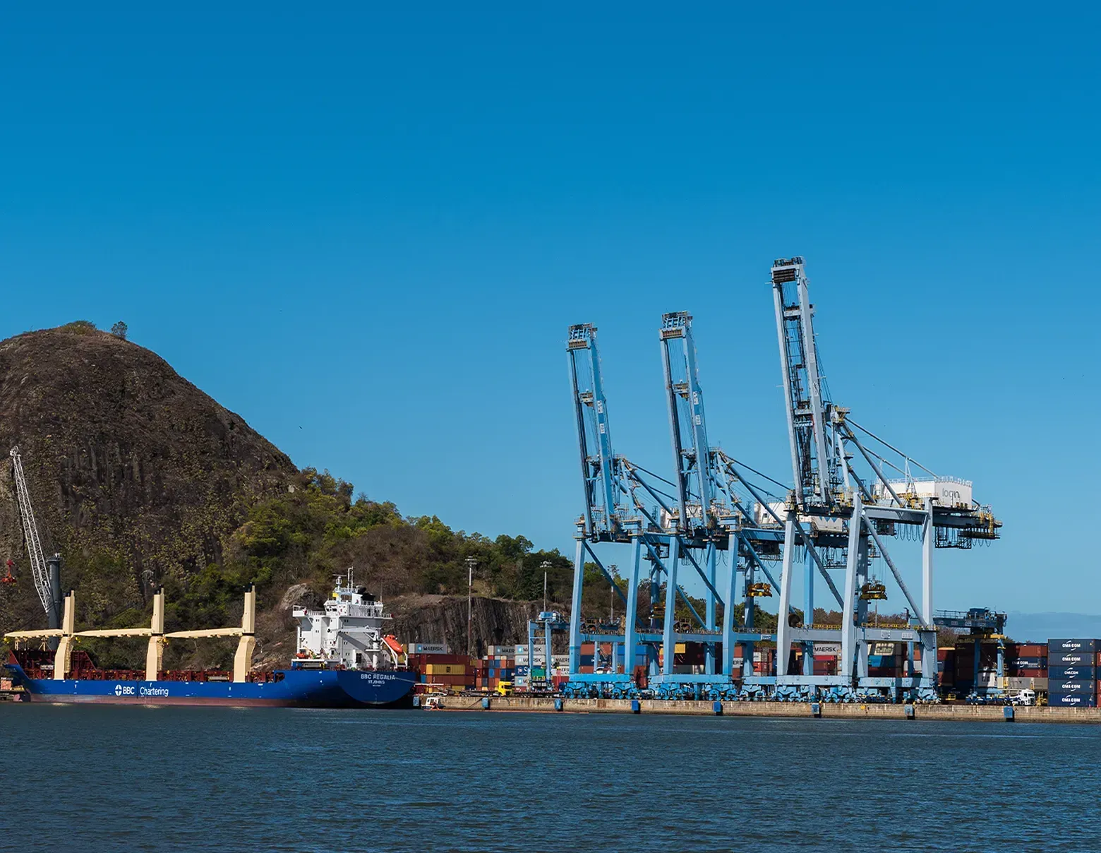 A large cargo ship docked at a port with three large cranes against a clear blue sky and a forested hill in the background.