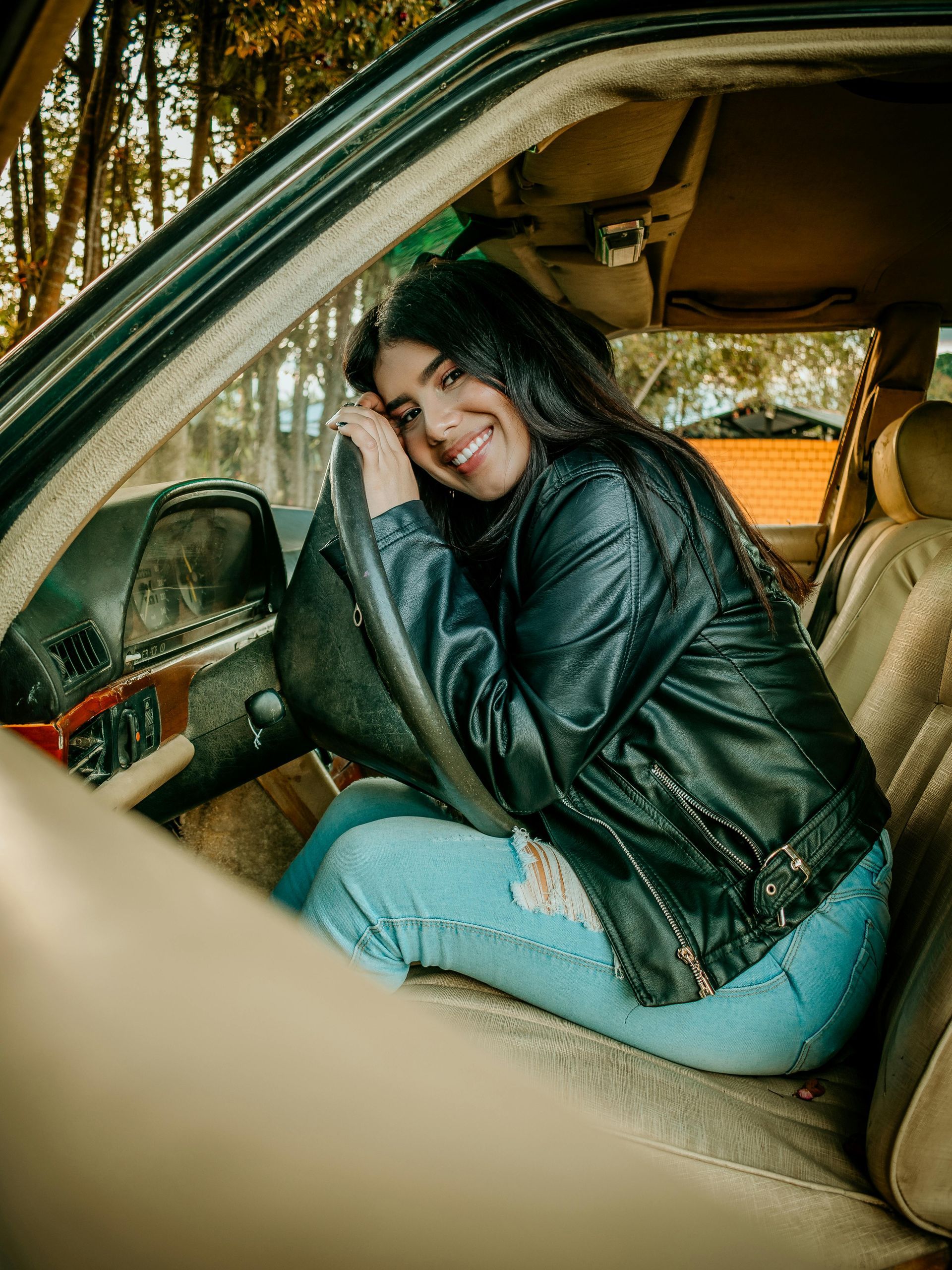 A woman in a leather jacket is sitting in the driver 's seat of a car.