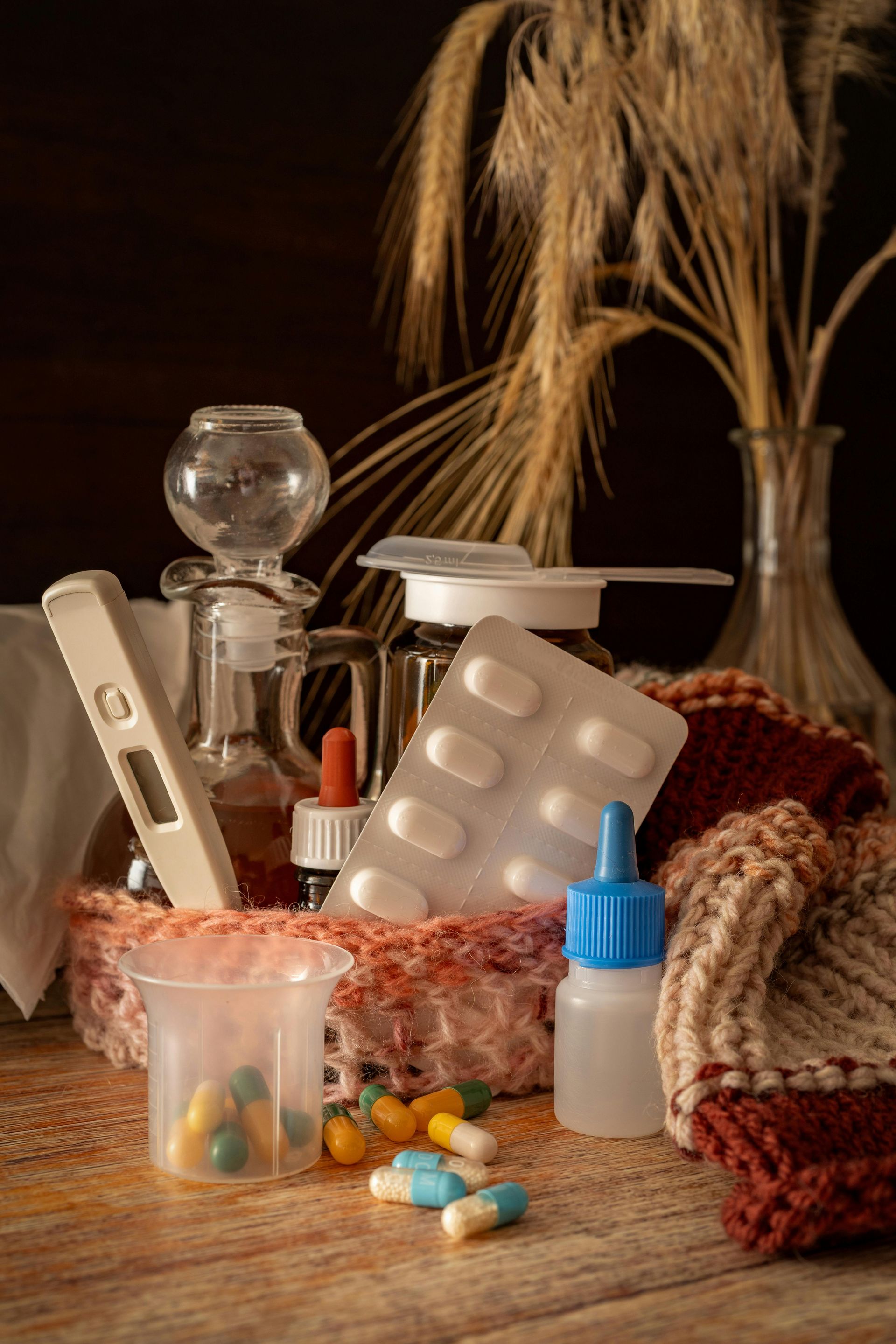 Assorted medicines and pill bottles on a table beside a vase with dried flowers and a knitted cloth