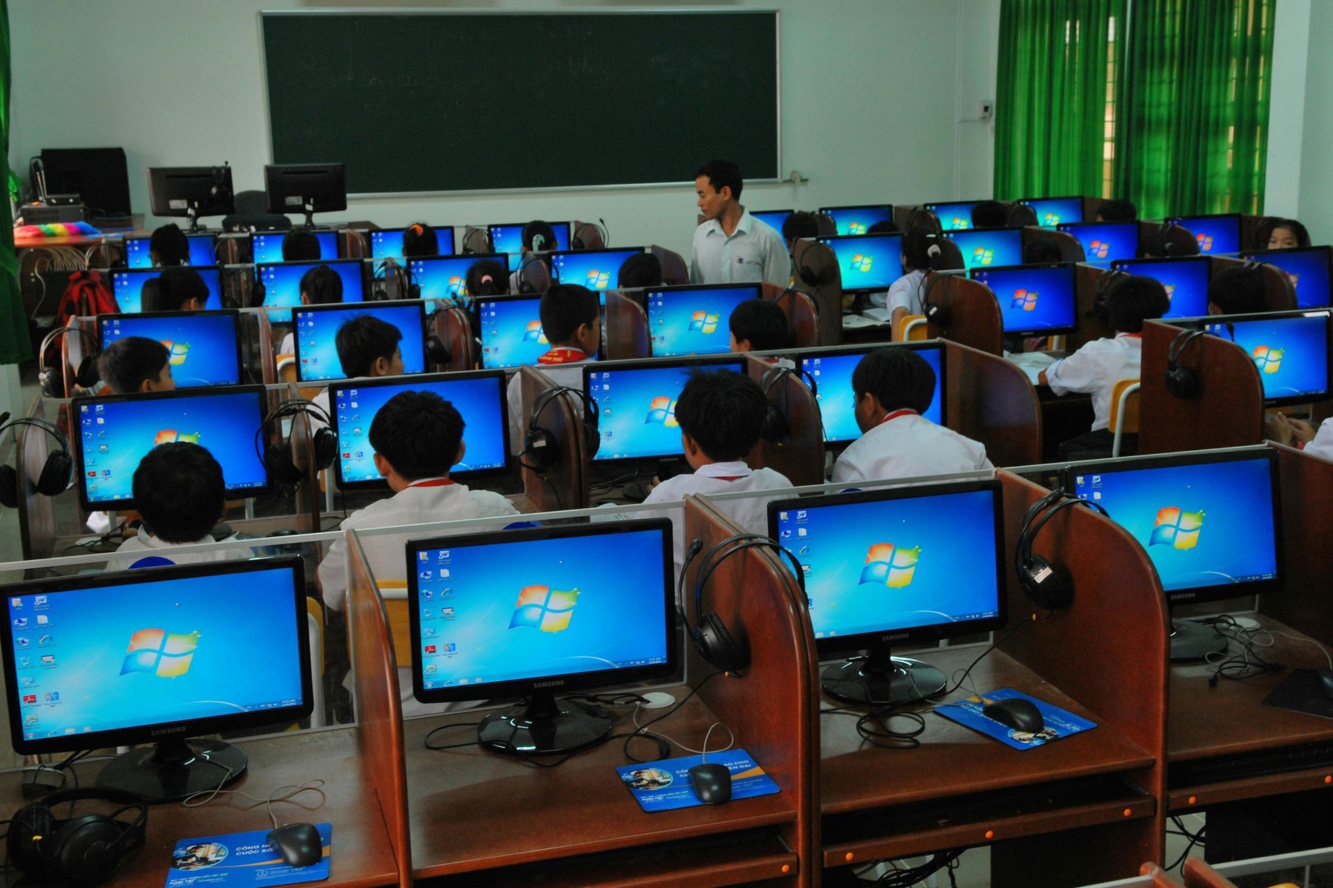 Computer lab with students at desktop PCs and a teacher at the front near a blackboard