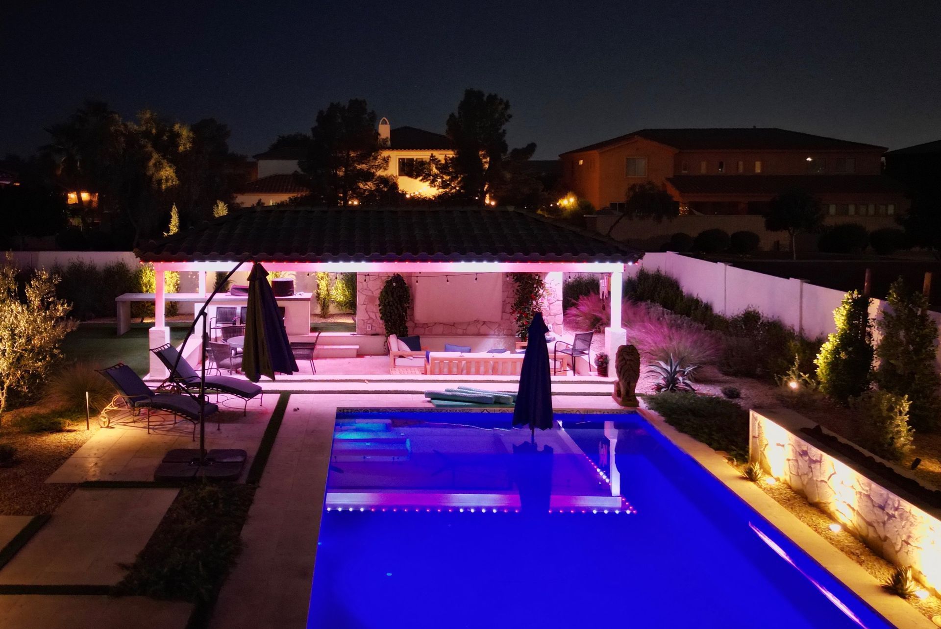 Pool area at night illuminated with colorful lights. Features a gazebo, lounge chairs, and landscaping.