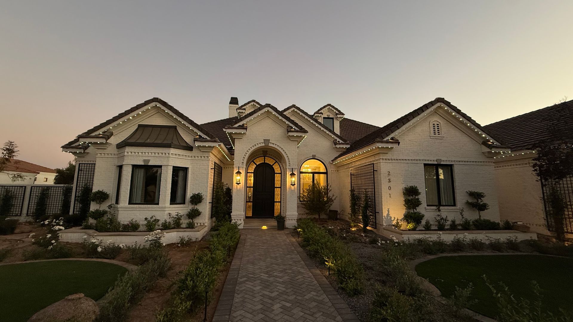Elegant stone house with arched entry, lit windows, and a landscaped front yard at dusk