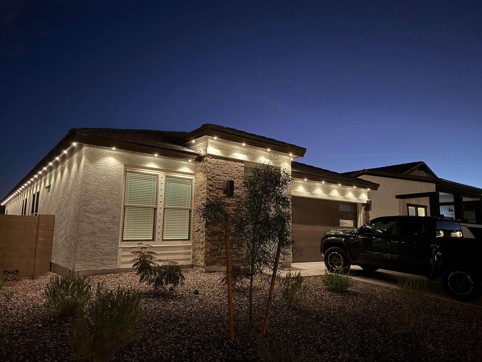 House at dusk with warm roofline lights, front yard shrubs, and a parked black truck in the driveway
