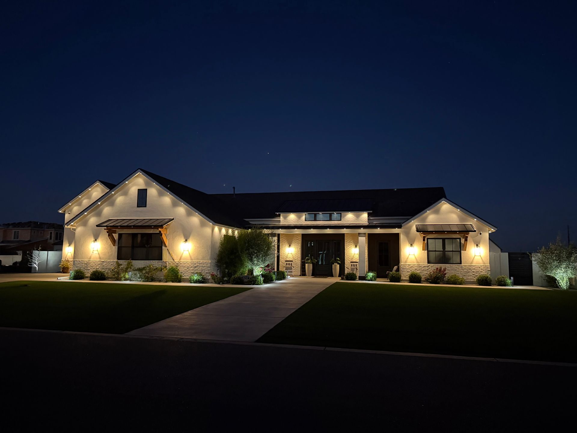 Modern stone house at dusk with warm porch lights and a dark landscaped lawn
