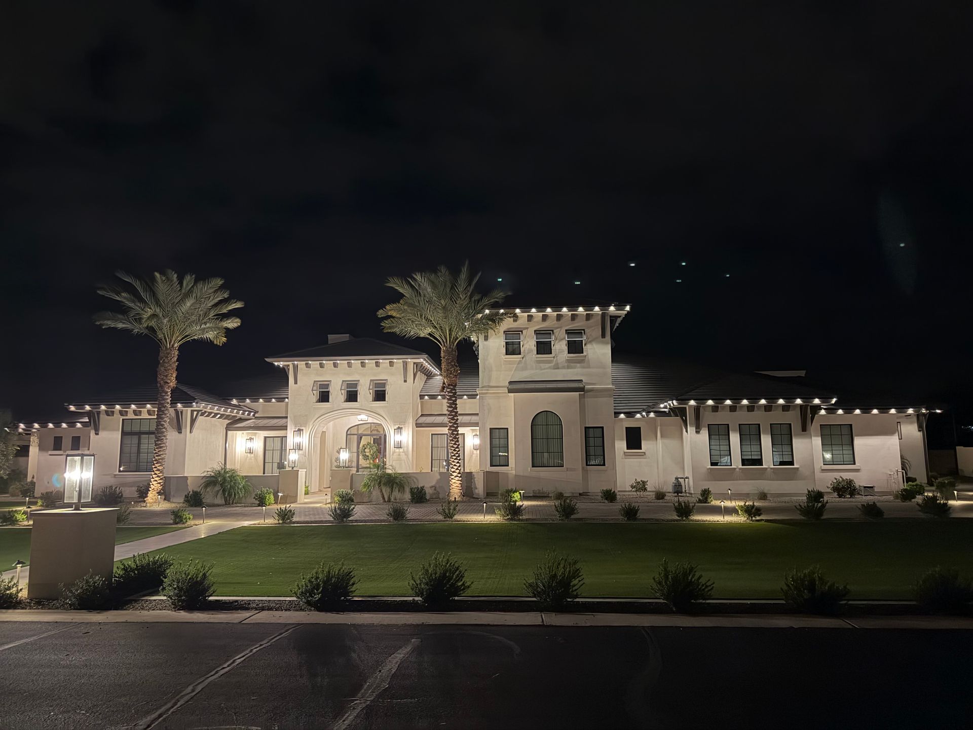 Nighttime view of a lit Mediterranean-style mansion with palm trees and landscaped gardens