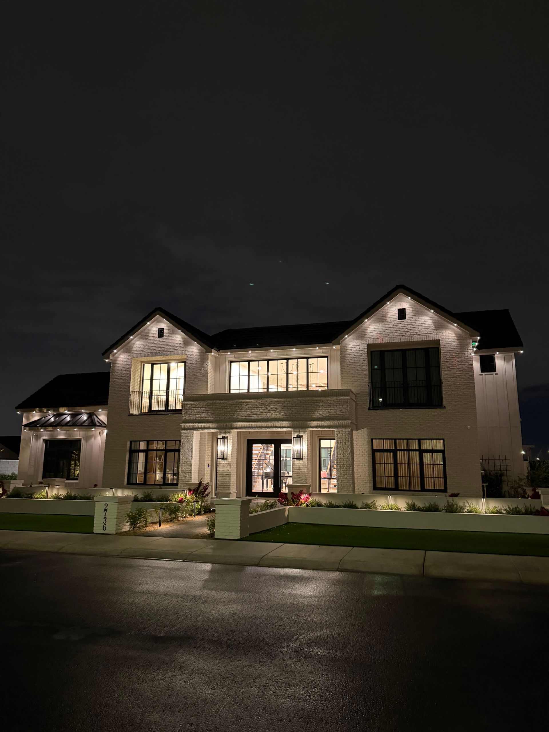 Large two-story stone house lit up at night, with a wet driveway and dark sky.