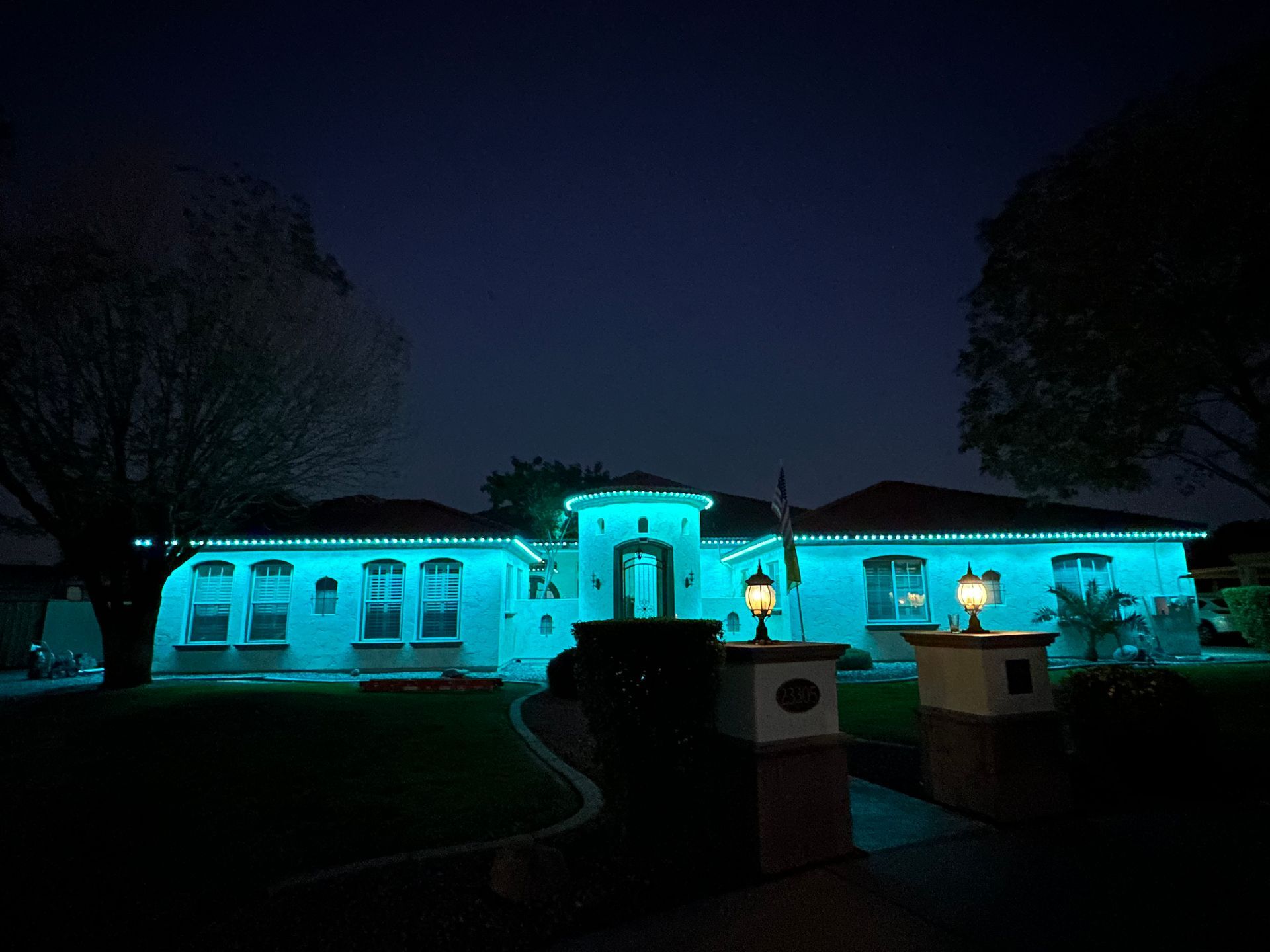 Night view of a turquoise-lit building with arched windows and a central entrance, framed by dark trees and lawn