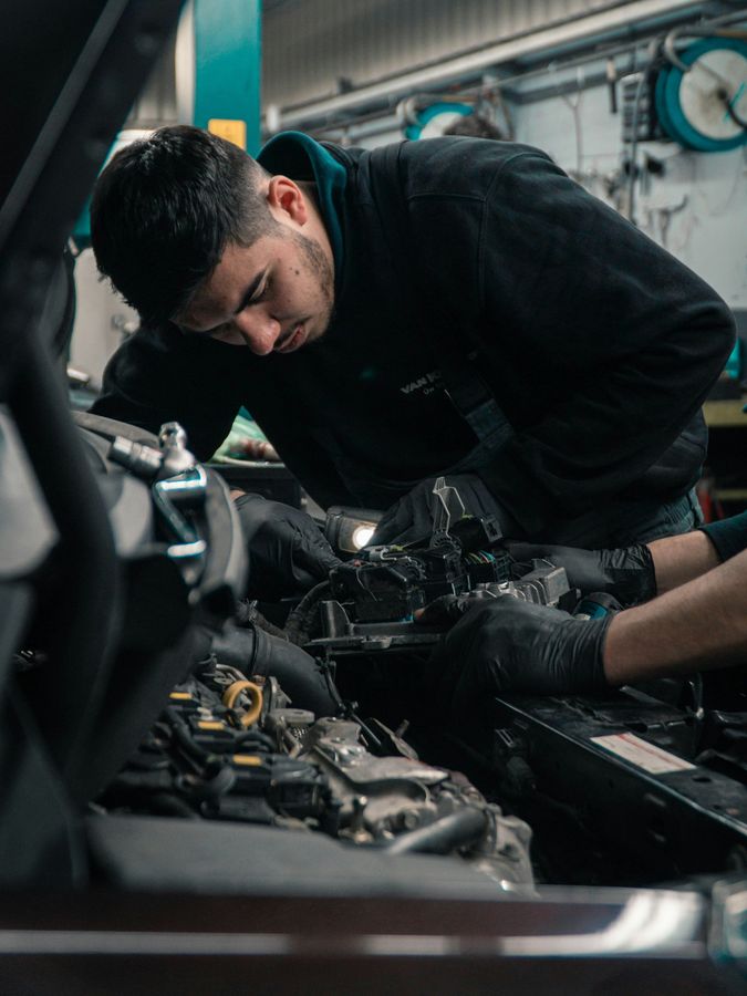 Mechanic in black clothing leaning over a car engine, working under the hood in a garage.