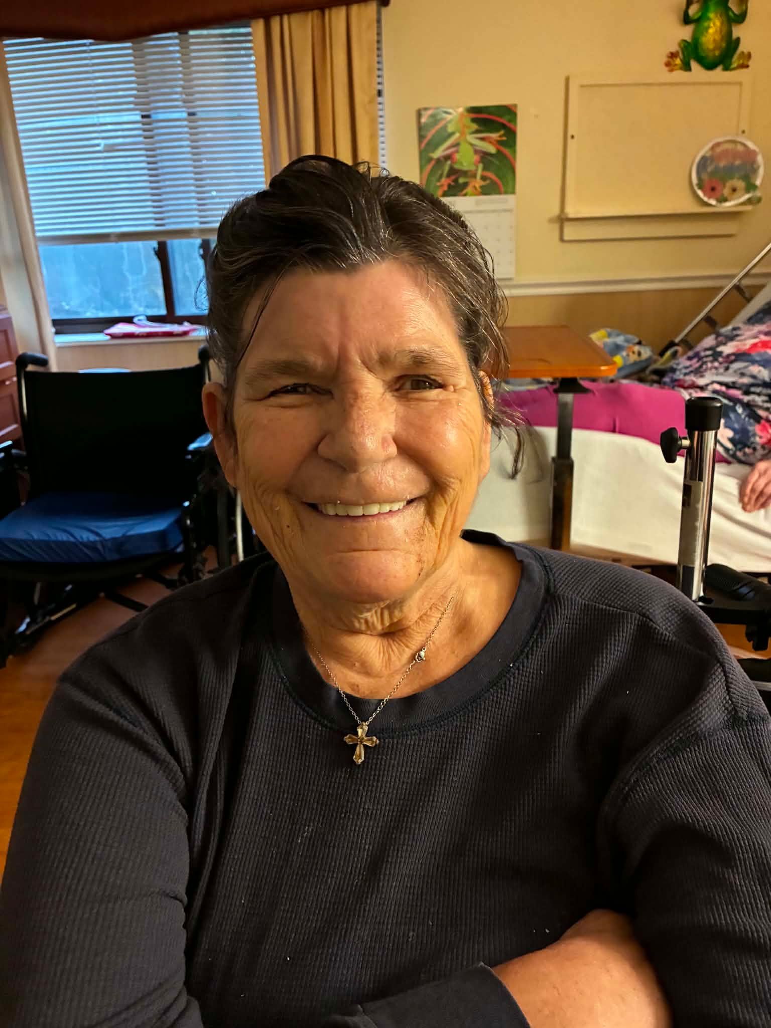 Smiling woman seated indoors by a window, wearing a dark top and necklace.