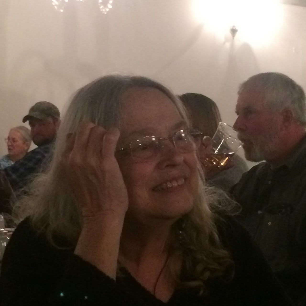 A woman wearing glasses is smiling while sitting at a table with other people