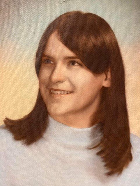 A woman with long brown hair is wearing a white turtleneck and smiling.