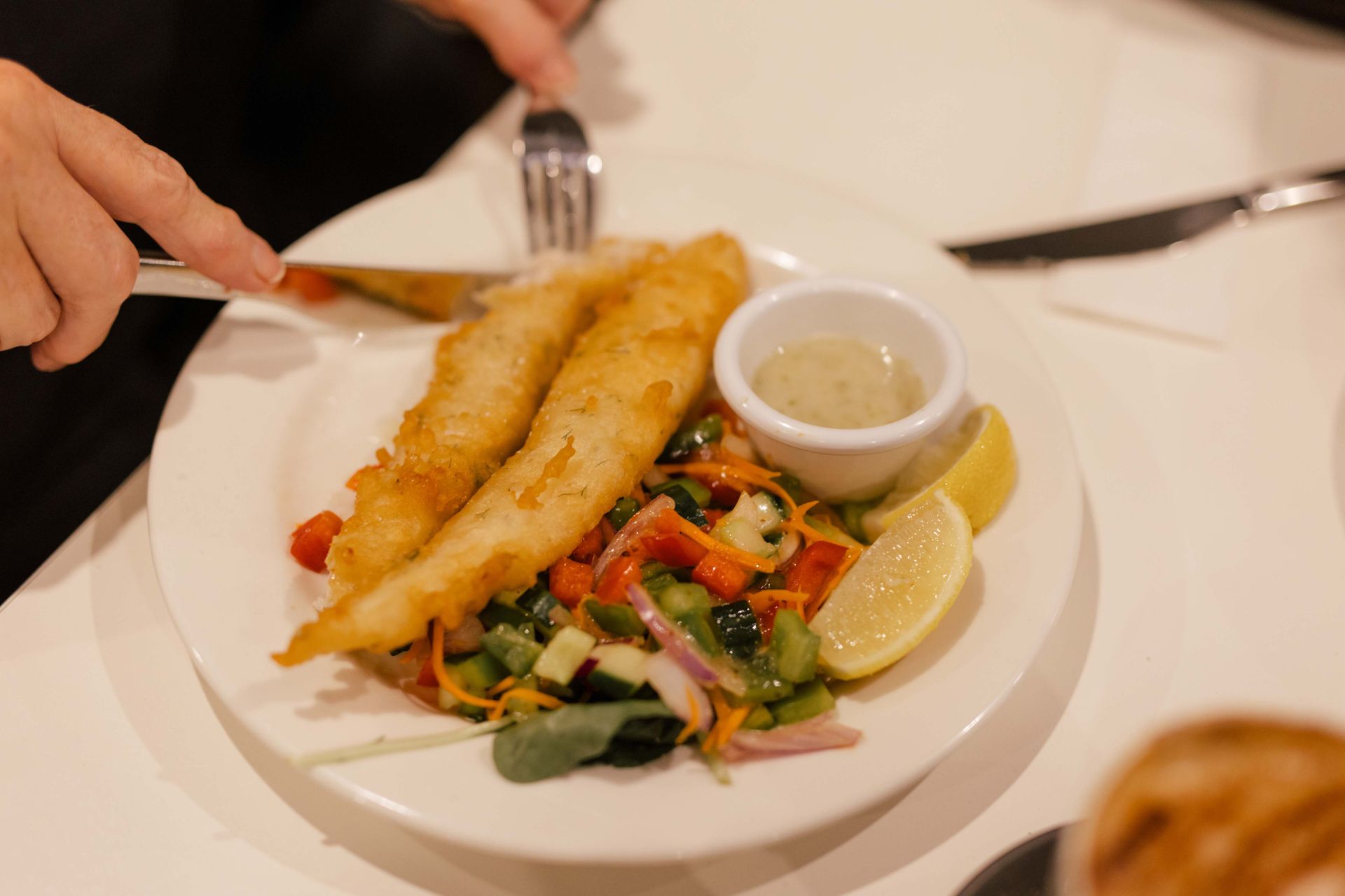 A person is cutting into a piece of fish on a plate with lemon, sauce and vegetables